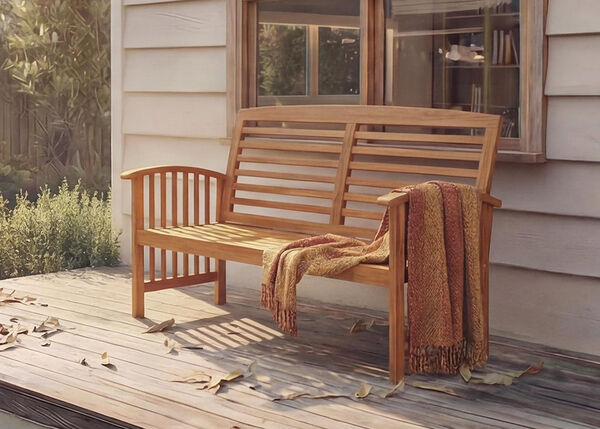 Wooden bench with a throw blanket on a deck with fallen leaves, beside a house.