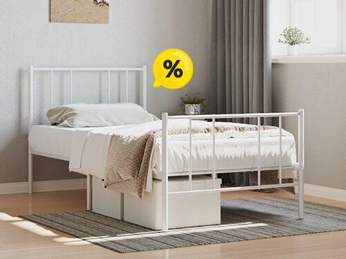An image showing a white metal bed with a white comforter and a textured throw blanket. A small potted plant sits on a nightstand next to the bed, and a yellow speech bubble containing a percentage symbol (%) floats above the bed. The bed is on a woven rug and a wall with light-colored wood paneling is partially visible in the background. Click to see the products in the collection.