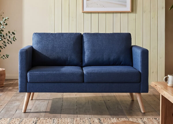 A navy blue sofa with light wood legs sits in a modern living room scene, alongside a potted plant and a wooden table with a mug. Above the sofa, a framed photograph shows a beach. Click to see the products in the collection