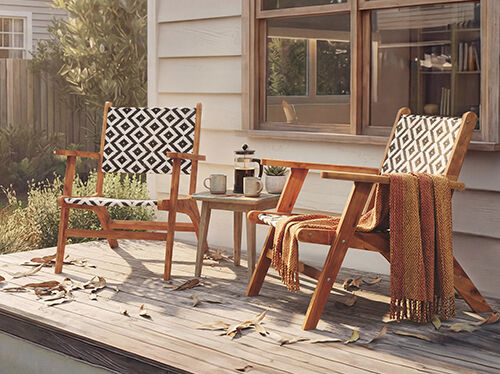 &nbsp;A wooden patio features two Adirondack chairs, a small side table, and fall foliage. One chair has a black and white patterned cushion, the other features a textured pattern, and a copper teapot sits next to two mugs on the table. A throw blanket is draped over one chair. Click to see the products in the collection.
