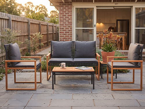 An outdoor patio setting featuring a gray sofa with two matching armchairs and a central coffee table with a wooden tabletop. The furniture has a modern, minimalist design with dark frames and light-colored cushions. A small potted plant adds a touch of greenery to the scene, and a mug sits on the coffee table. In the background, there is a brick facade with windows and a wooden fence. Click to see the products in the collection