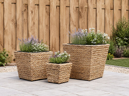 Three woven square planters with flowering plants arranged on a paved outdoor patio in front of a timber fence. Click to see the products in the collection.