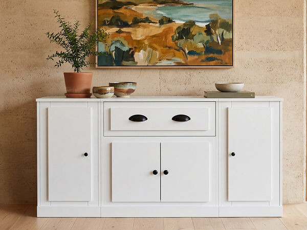 A white sideboard with black handles and drawers sits against a textured beige wall. Above the sideboard hangs a framed painting with earthy tones, and to the right and left are potted plants in terracotta and neutral-colored containers. A wooden book rests on top of the sideboard. Click to see the products in the collection.