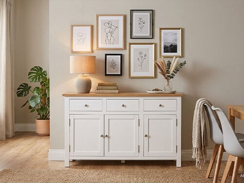 A light beige wall displays a collection of framed artwork in varying sizes and shapes, arranged above a white sideboard. A table lamp, potted plant, and vase with dried florals rest on the sideboard. Click to see the products in the collection.