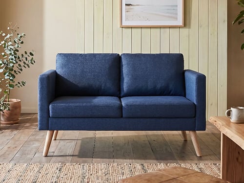 A navy blue sofa with light wood legs sits in a modern living room scene, alongside a potted plant and a wooden table with a mug. Above the sofa, a framed photograph shows a beach. Click to see the products in the collection