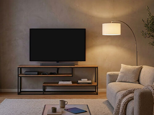A modern living room features a large television on a wooden console, illuminated by an arched floor lamp. A grey sofa and coffee table with a mug and tablet are also visible. Click to see the products in the collection.