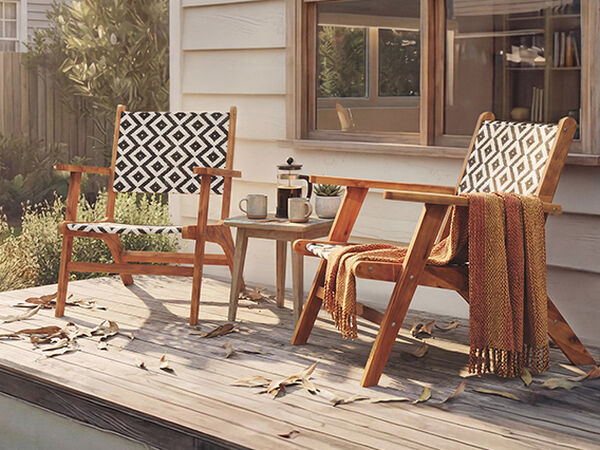 &nbsp;A wooden patio features two Adirondack chairs, a small side table, and fall foliage. One chair has a black and white patterned cushion, the other features a textured pattern, and a copper teapot sits next to two mugs on the table. A throw blanket is draped over one chair. Click to see the products in the collection.