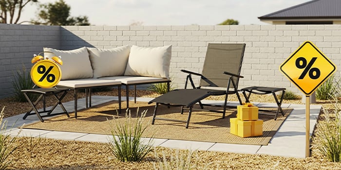 A grey outdoor lounge set with cream cushions, a matching sunlounger and side tables, yellow gift boxes, a sign, and a clock with discount icons, all on a styled patio. Click to shop the collection.